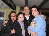 Family of four with a small dog indoors; smiling, embracing.