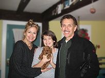 Family of three with a small brown dog indoors. Woman, girl, and man smile.