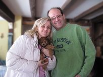 Couple smiling, woman holding small dog, man in green sweatshirt, indoors.