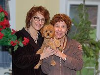 Two women with red hair holding a small, brown dog; smiling, near a decorated plant.
