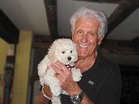 Smiling older man holding a small white puppy indoors. Man wears a black shirt and a watch.