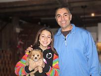 Girl and man holding a small, tan puppy. Girl smiles, wearing colorful jacket; man in blue shirt, both indoors.