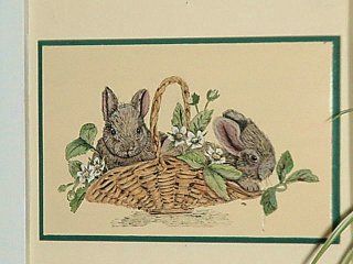 Two brown bunnies peek over a wicker basket filled with white flowers and green leaves; cream background, green border.