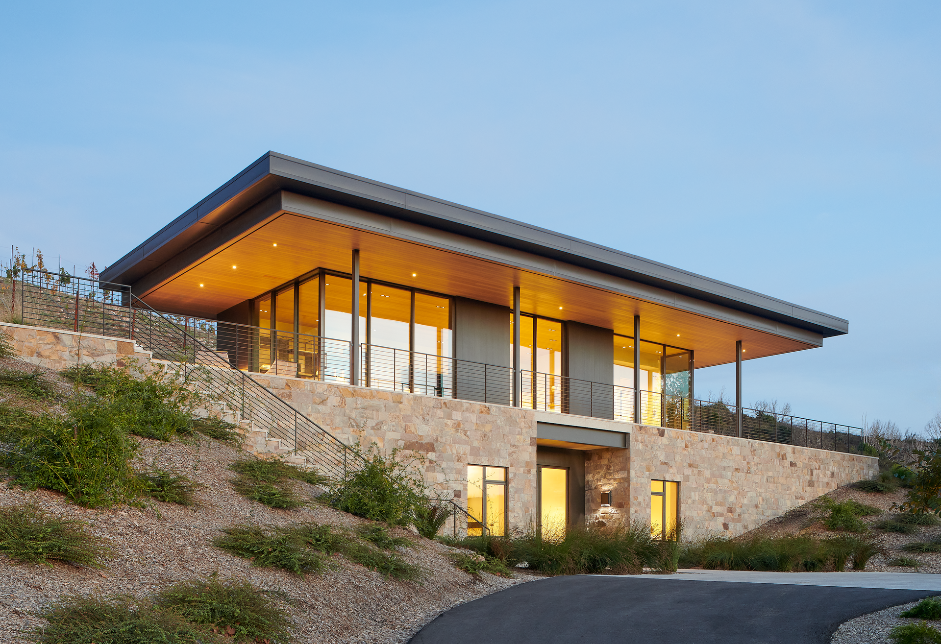 Modern building nestled into hillside, stone walls, glass windows, flat roof, evening light.