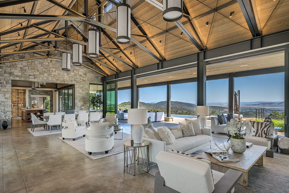 Spacious living room with stone walls, vaulted wood ceiling, and large windows overlooking a scenic landscape.