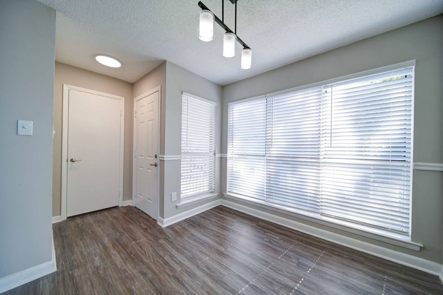 An empty living room with hardwood floors , blinds and a large window.