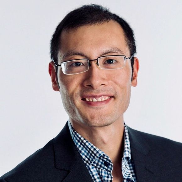 Man wearing glasses, smiling, with a checkered shirt and dark blazer against a white backdrop.