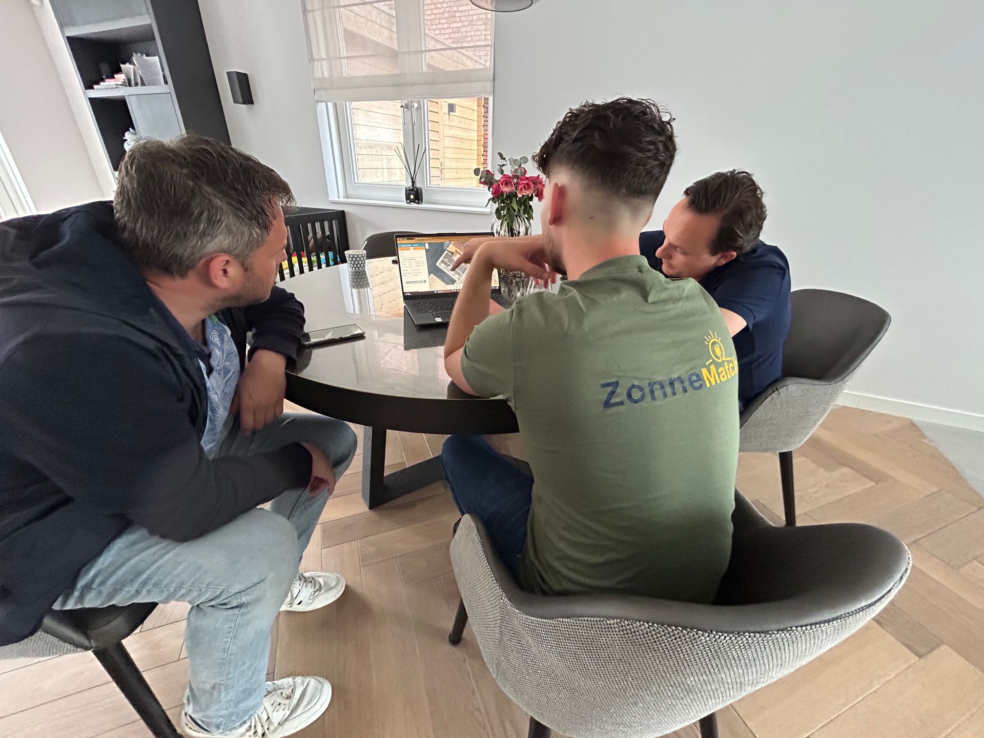 Drie mannen in vrijetijdskleding zitten rond een tafel, wijst er een aan, en bespreken iets binnenshuis.