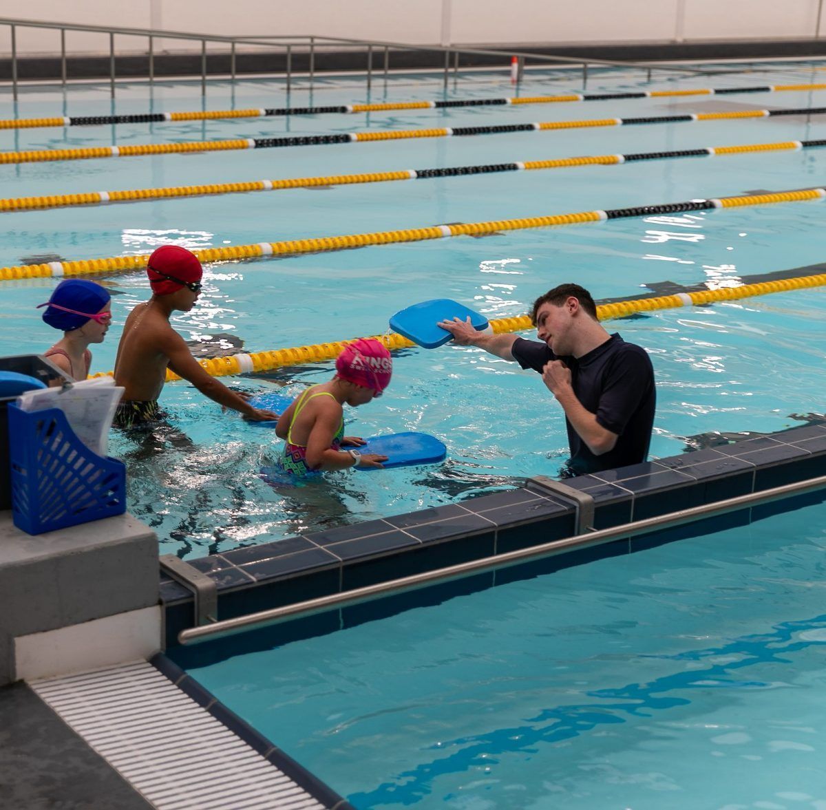A group of children are swimming in a pool with a man talking to them