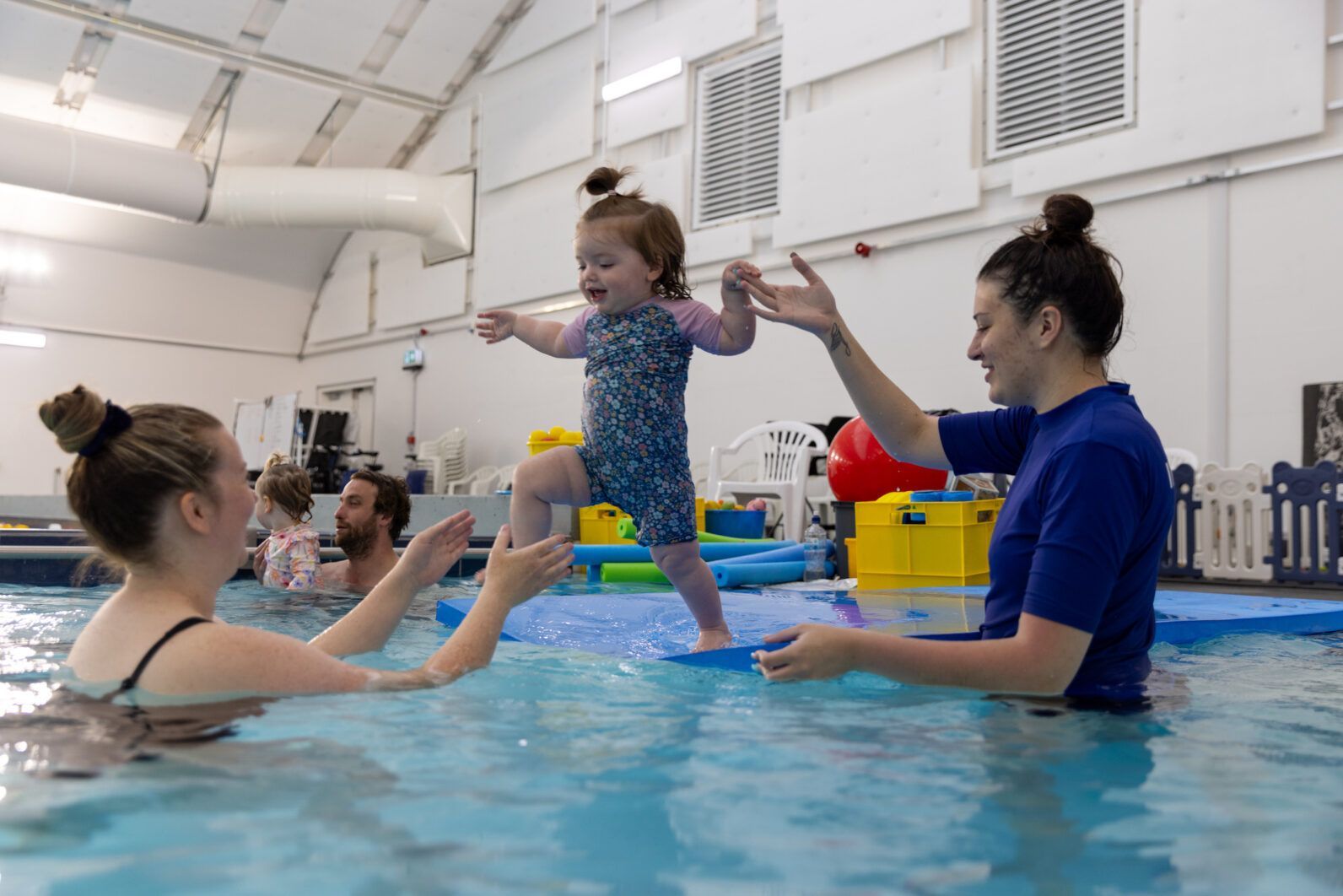 A little girl is standing on a raft in a swimming pool.