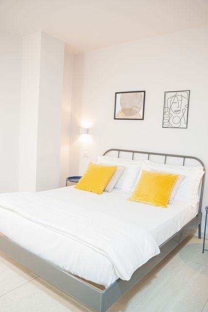 Camera da letto luminosa e minimalista con letto bianco, cuscini gialli e quadri appesi alle pareti.