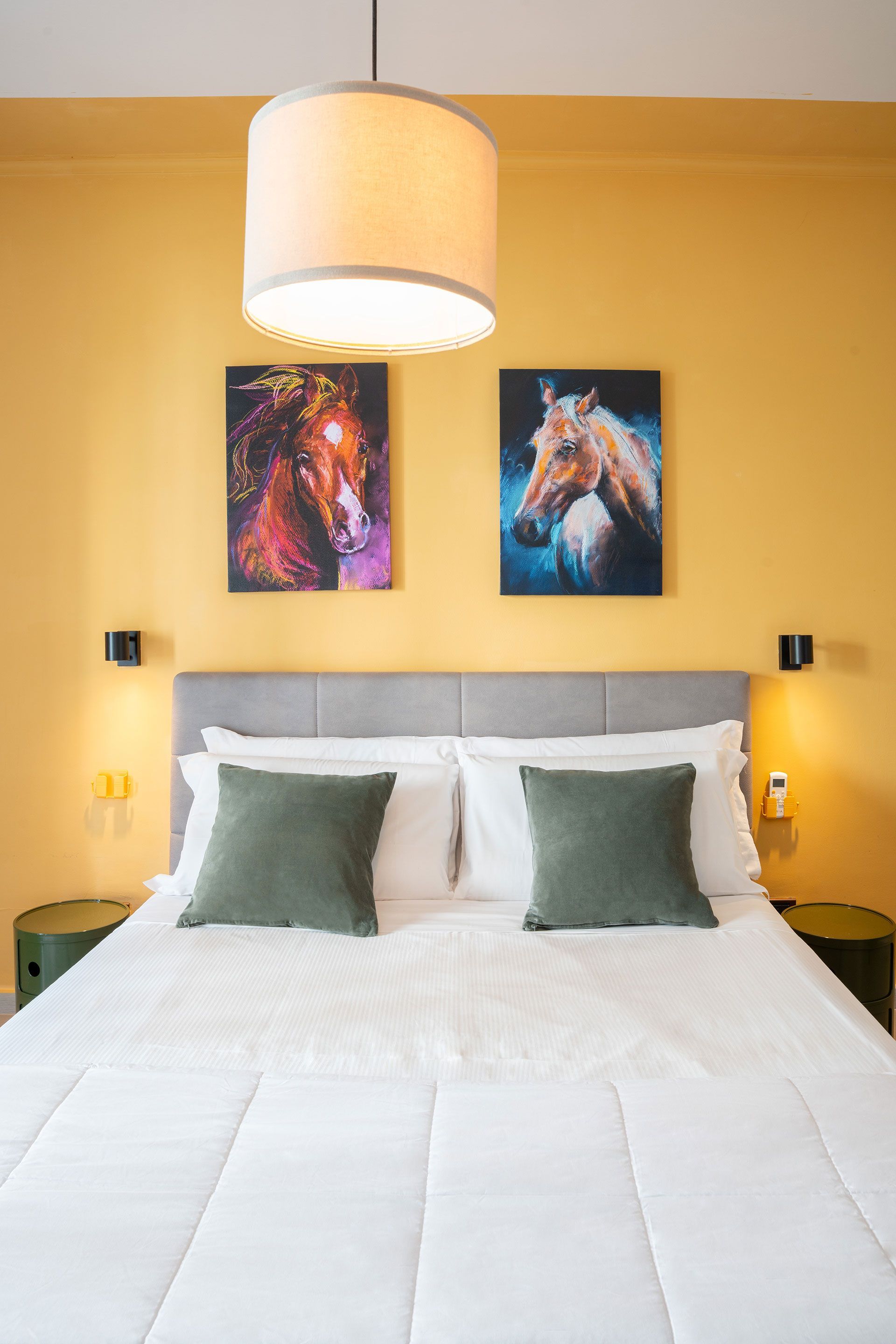 Un letto ben rifatto in una camera da letto gialla con colorati quadri raffiguranti cavalli e una lampada a sospensione.