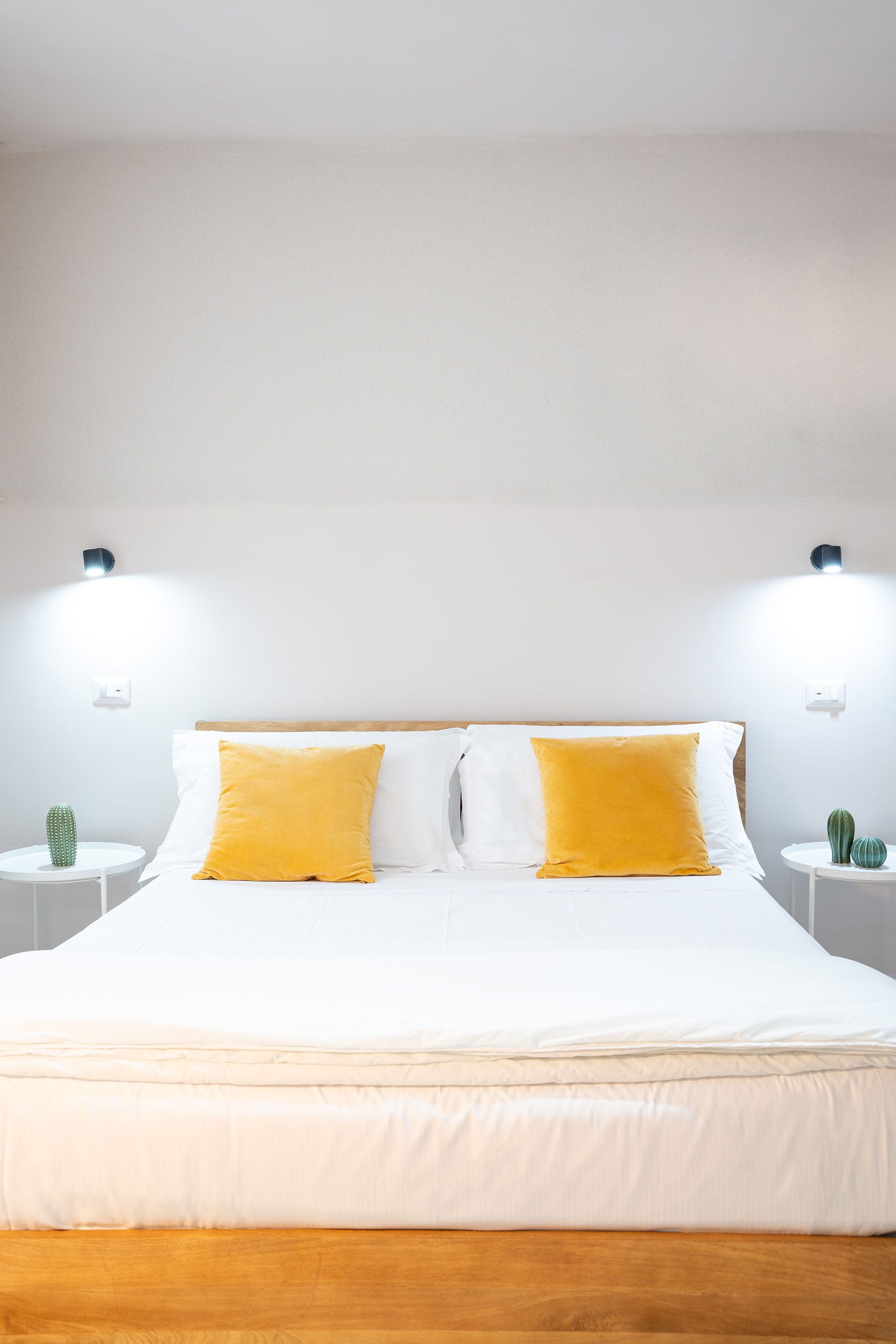 Luminosa camera da letto minimalista con un grande letto bianco e due cuscini gialli sotto lampade a parete.