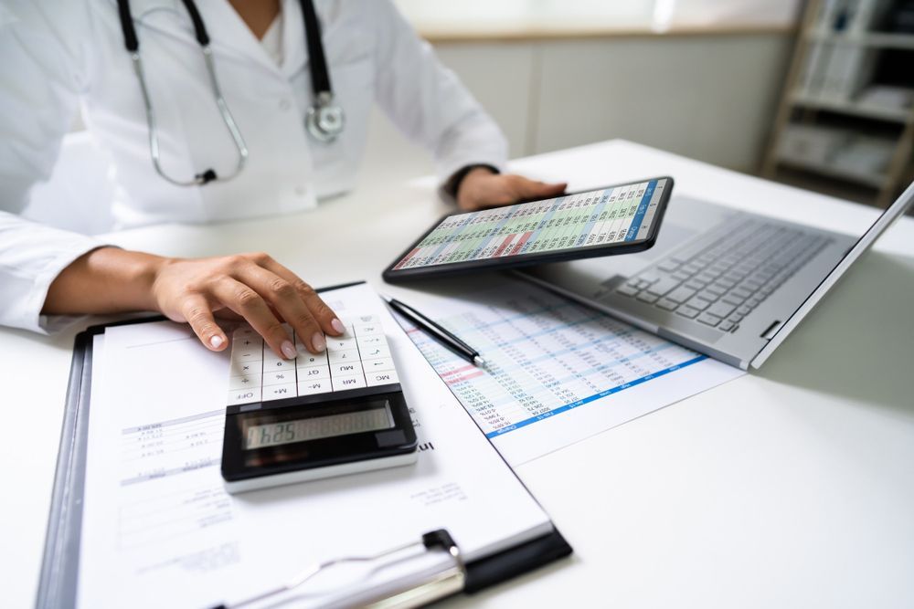 A doctor is sitting at a desk using a calculator and a tablet.