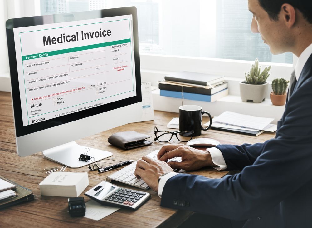 A man is sitting at a desk in front of a computer looking at a medical invoice.