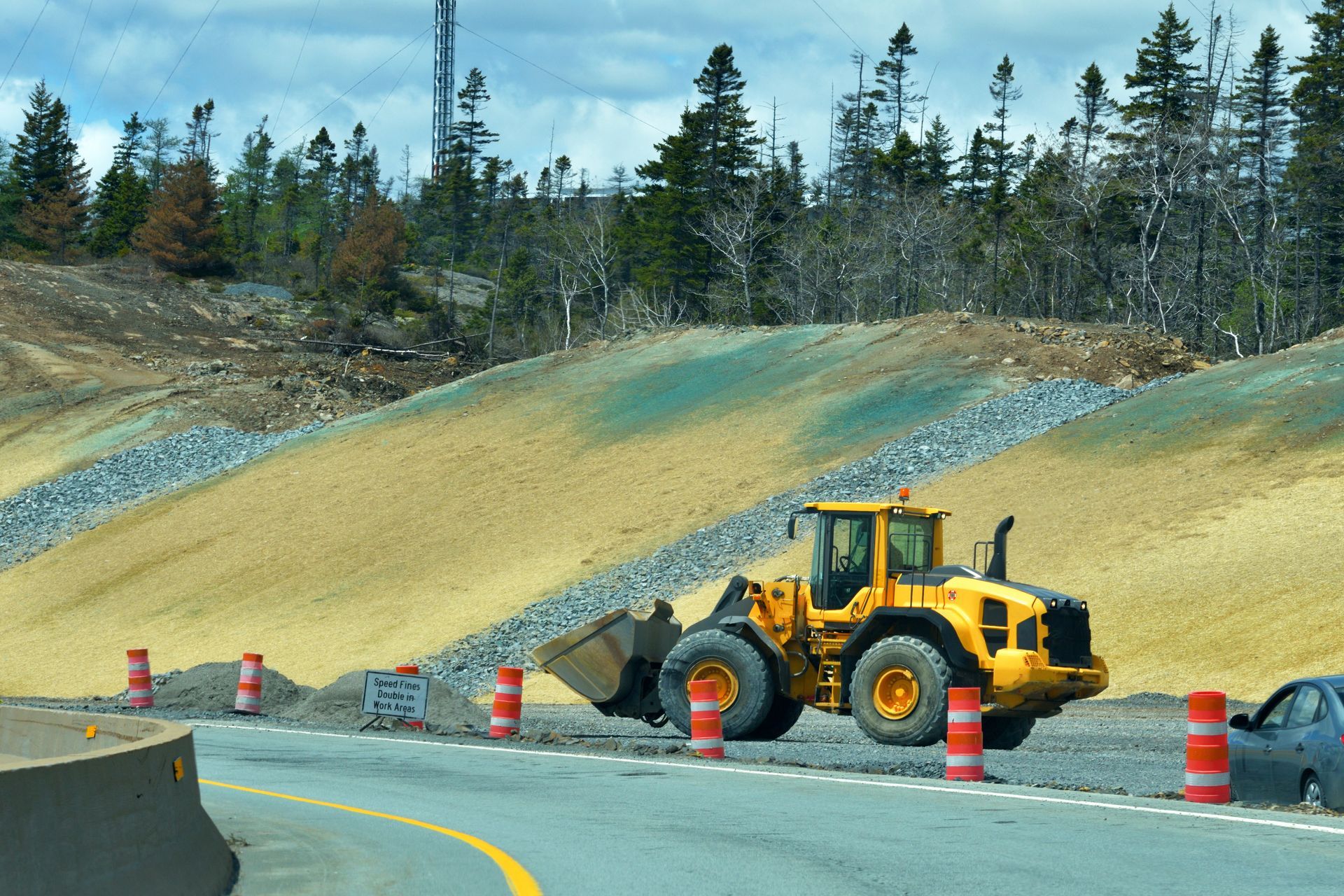 Yellow front loader on a road beside a sloped dirt embankment with orange cones and trees in the background