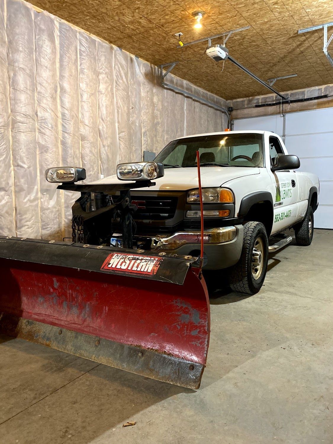 A white truck with a snow plow attached to it is parked in a garage.