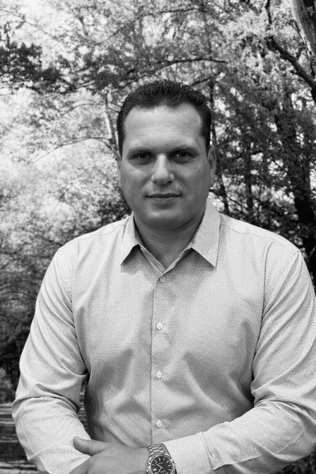 A person with short dark hair in a light-colored, button-down patterned shirt, standing in a wooded area.