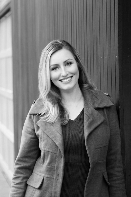 Smiling person wearing a coat, leaning against a paneled wall in a black-and-white photo.