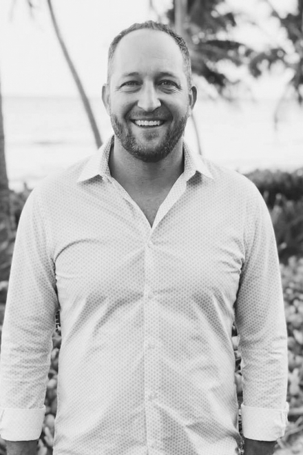 A man in a collared shirt smiling outdoors with tropical trees in the background.