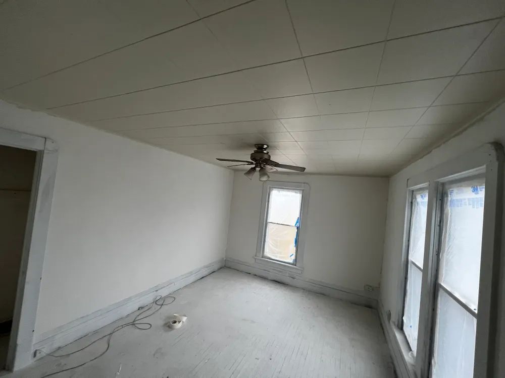 An empty room with a ceiling fan and two windows.