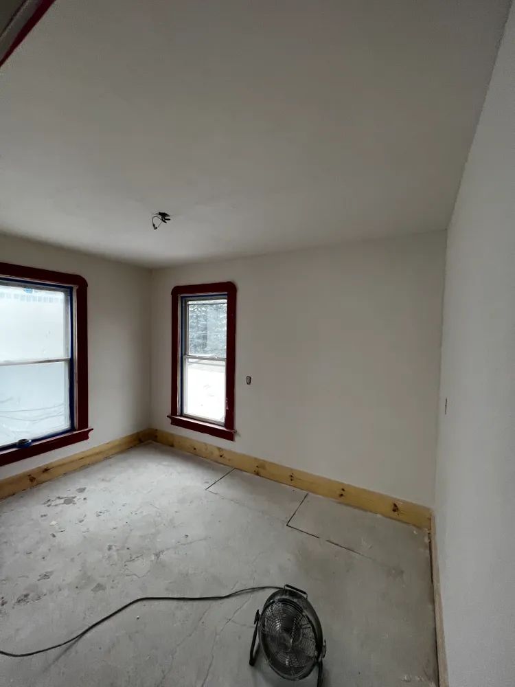 An empty room with two windows and a fan on the floor.