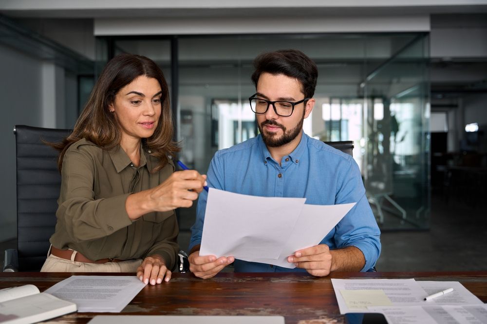 Two people in a modern office looking at documents on a desk, one pointing while the other holds the papers.