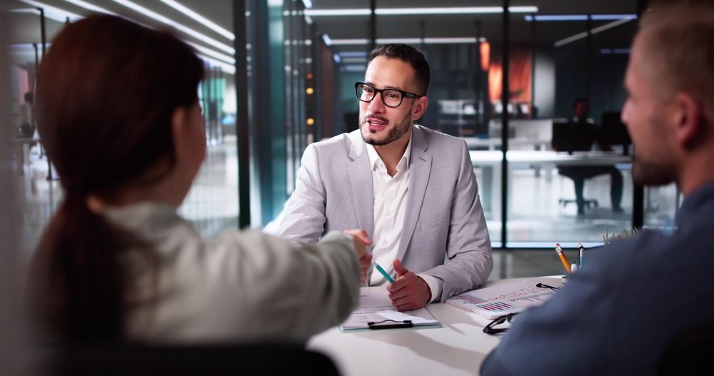 Two people in a meeting while a man in glasses speaks at a desk in a glass-walled office