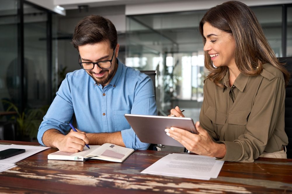 Two colleagues collaborate at a wooden desk; one writes in a notebook while the other holds a tablet.