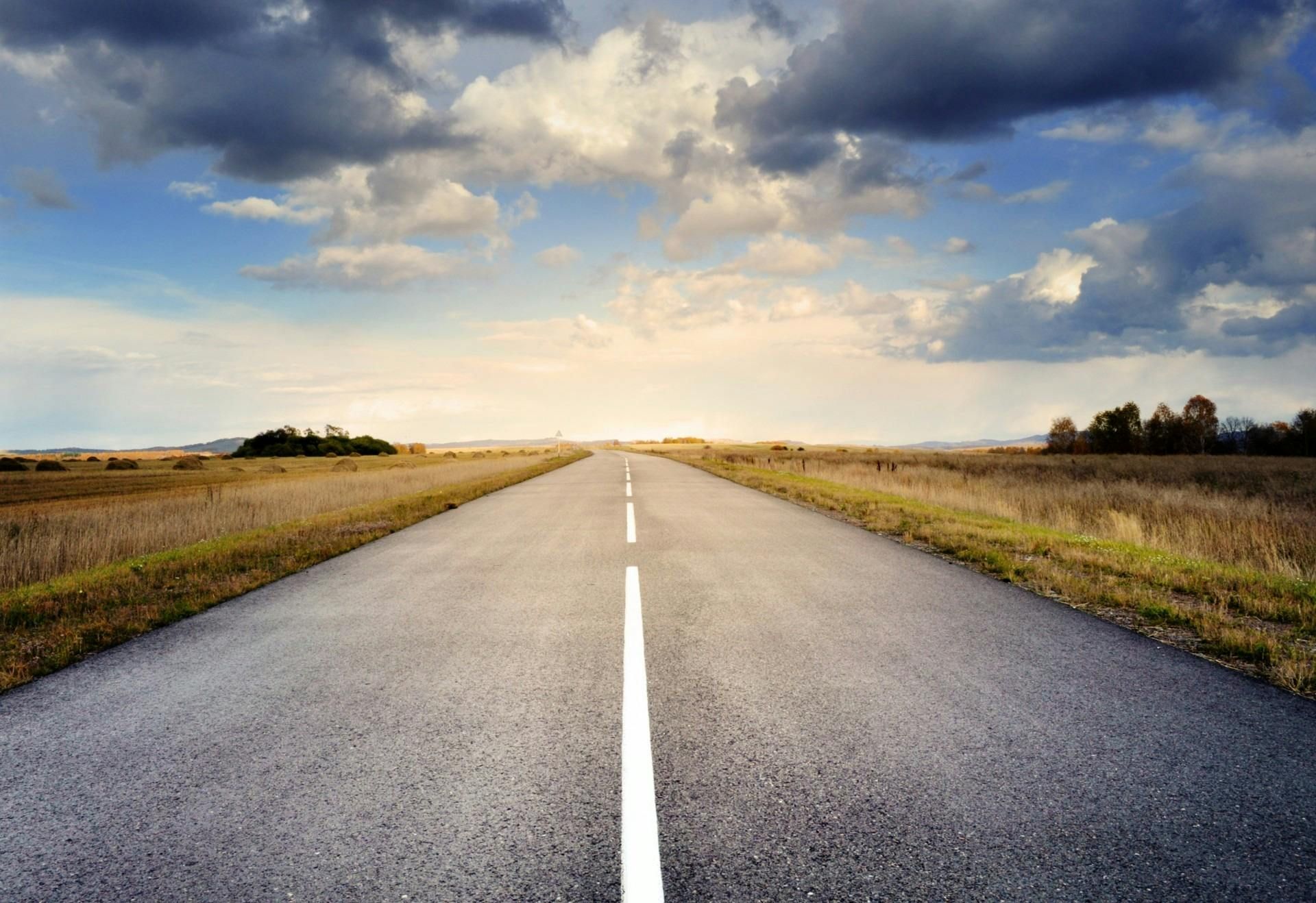 Road stretching towards the horizon under a dramatic sky with clouds, flanked by fields and trees.