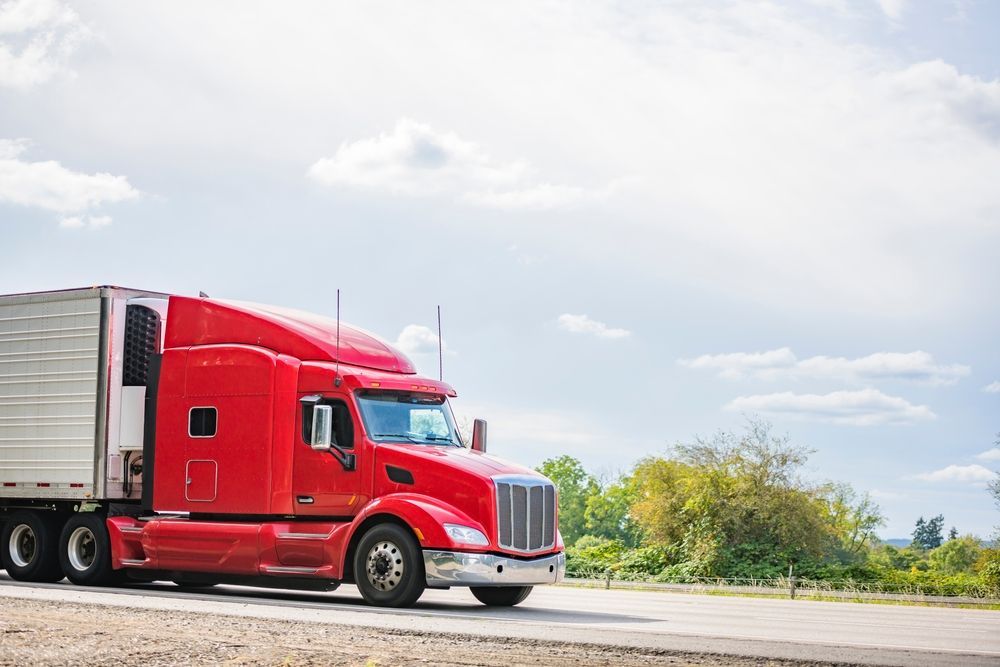 Red semi-truck driving on a road, cloudy sky background.