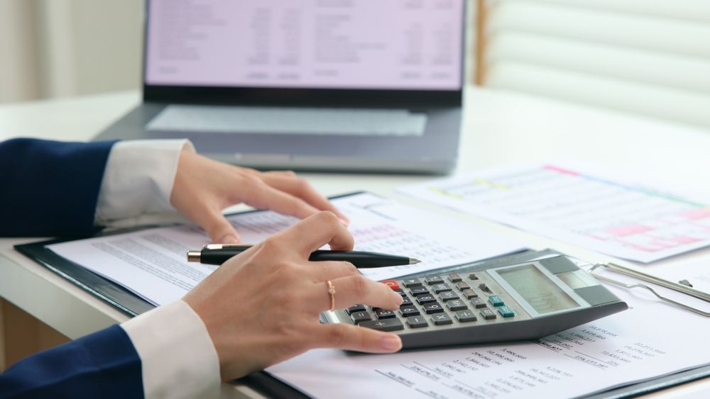 A person is using a calculator at a desk in front of a laptop.