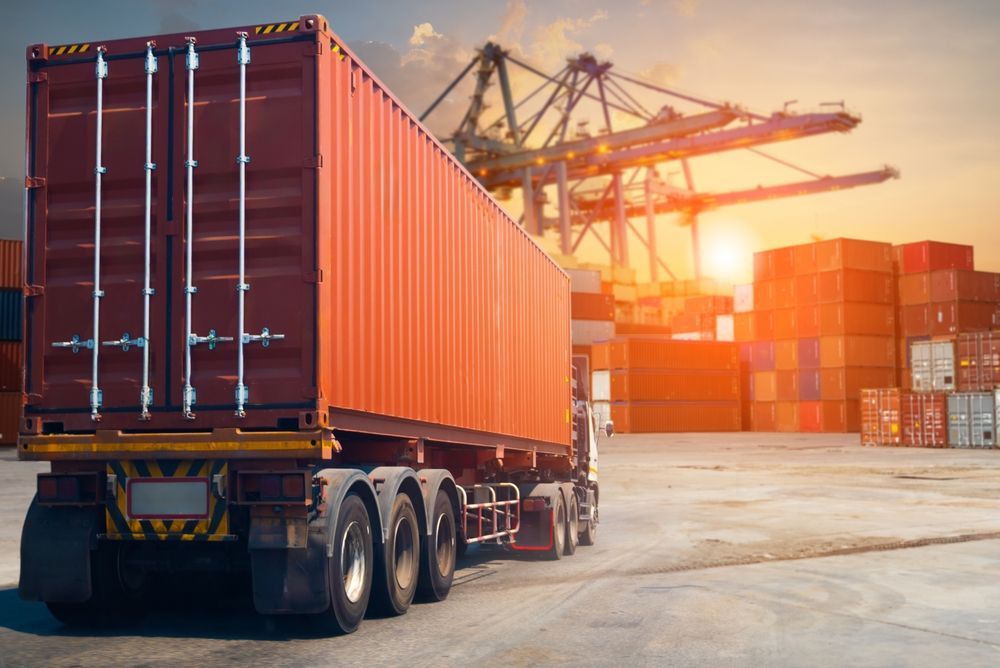 A semi truck is carrying a shipping container in a port at sunset.