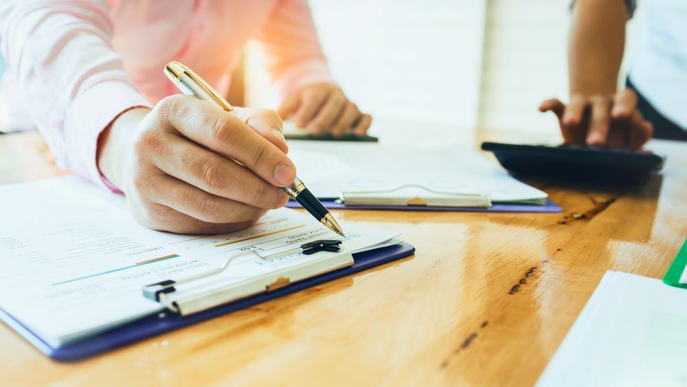 A person is writing on a clipboard while another person is using a calculator.