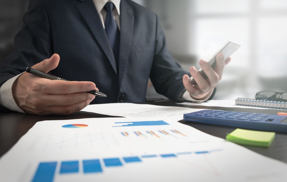 Person in a suit reviews financial charts with a calculator and phone on a desk.