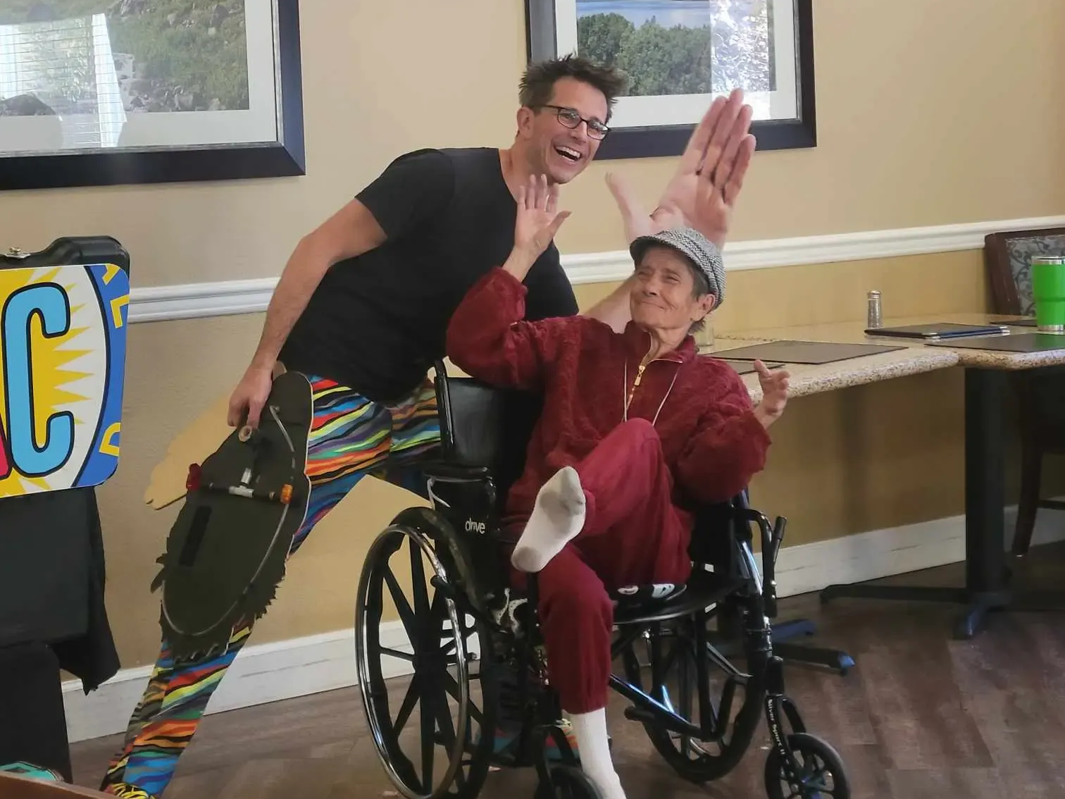 Man and person in wheelchair pose with a large hand-shaped hat. The man wears colorful pants and smiles.