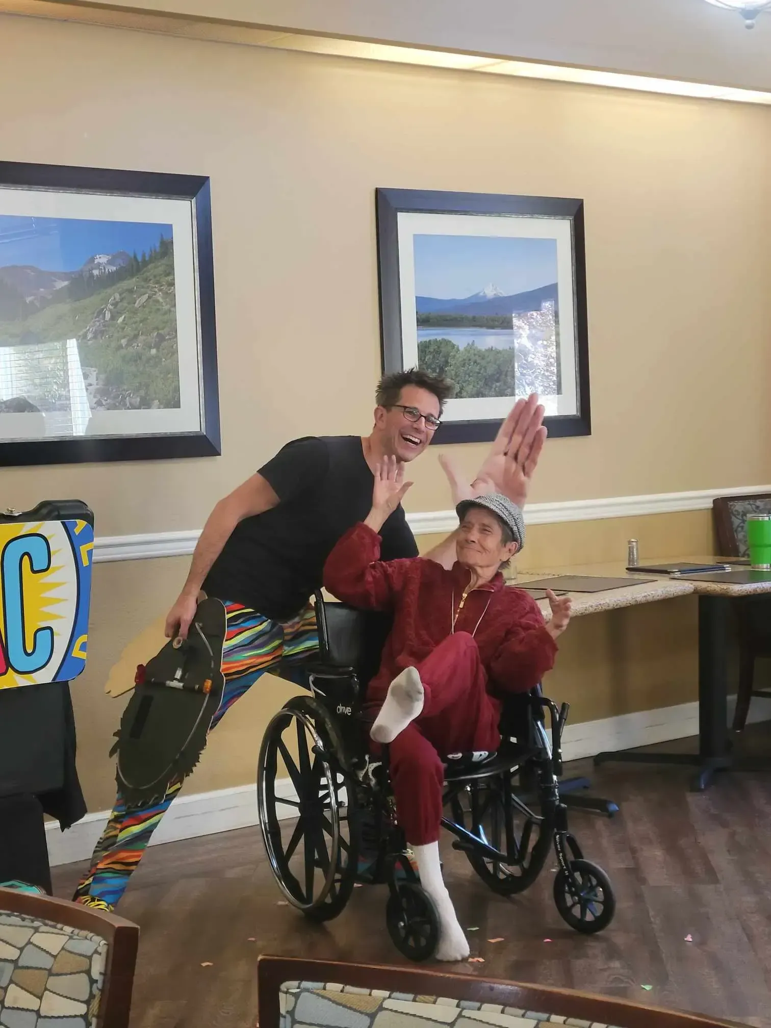 Man with colorful pants and woman in wheelchair posing with a tall hat; both smiling and gesturing, room with framed art.