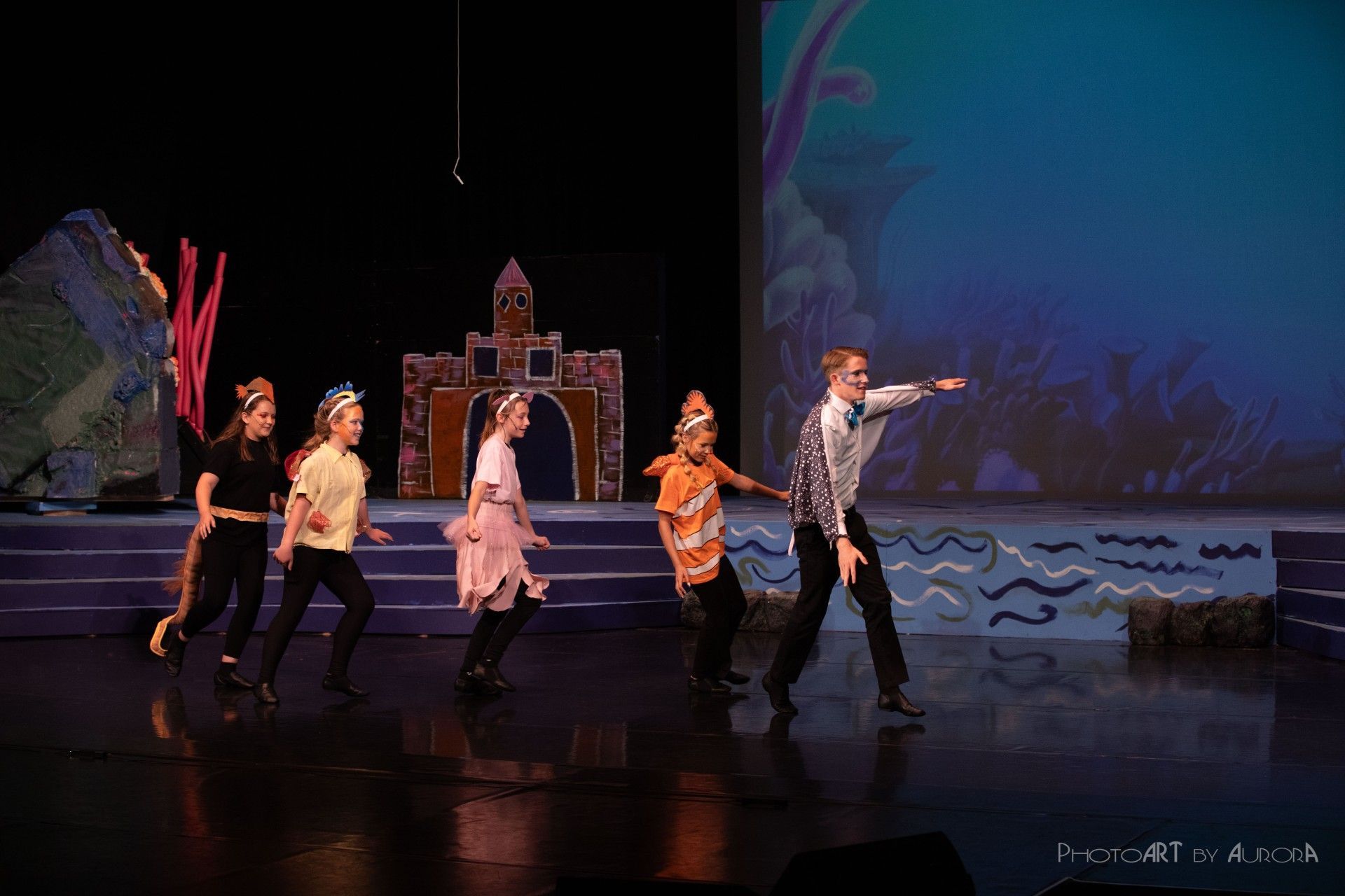 Actors on stage perform, dressed as ocean creatures, with a backdrop of an underwater scene.