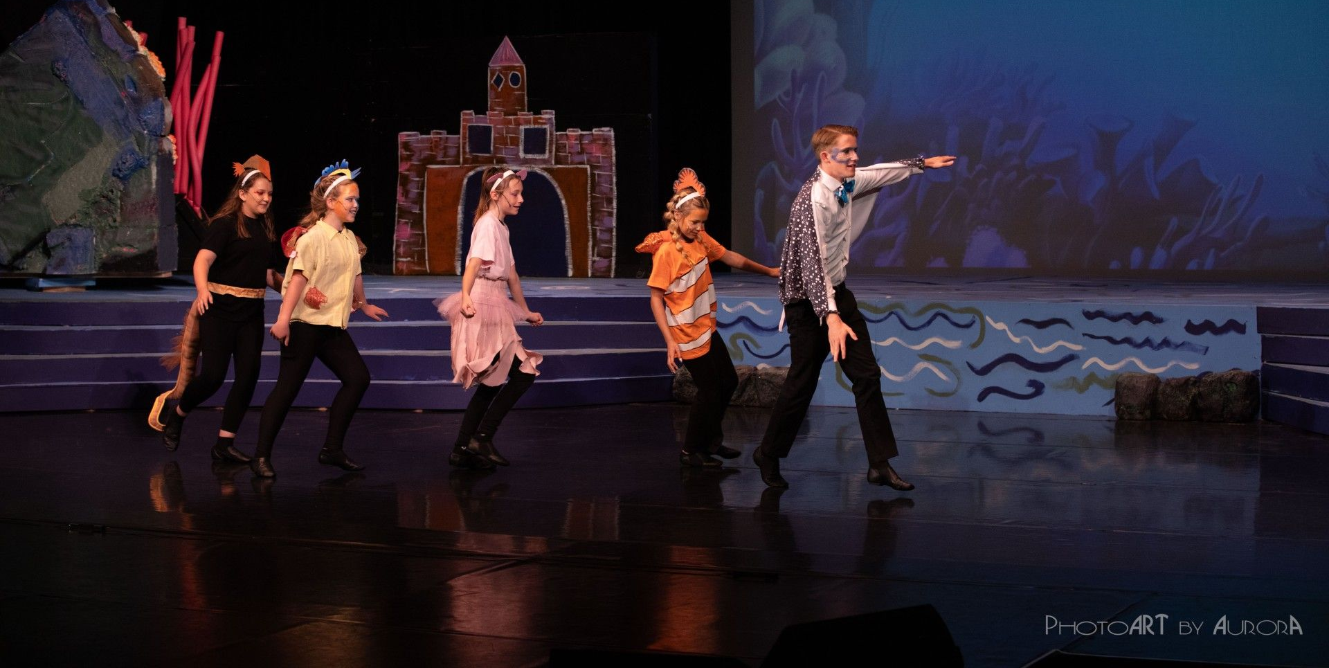 Children in costumes on stage, dancing in a play. Blue backdrop, castle prop.