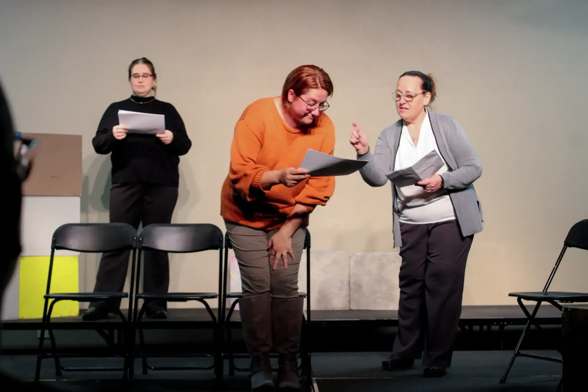 Three people on a stage rehearsing. One points, another bows, and the third stands holding papers.