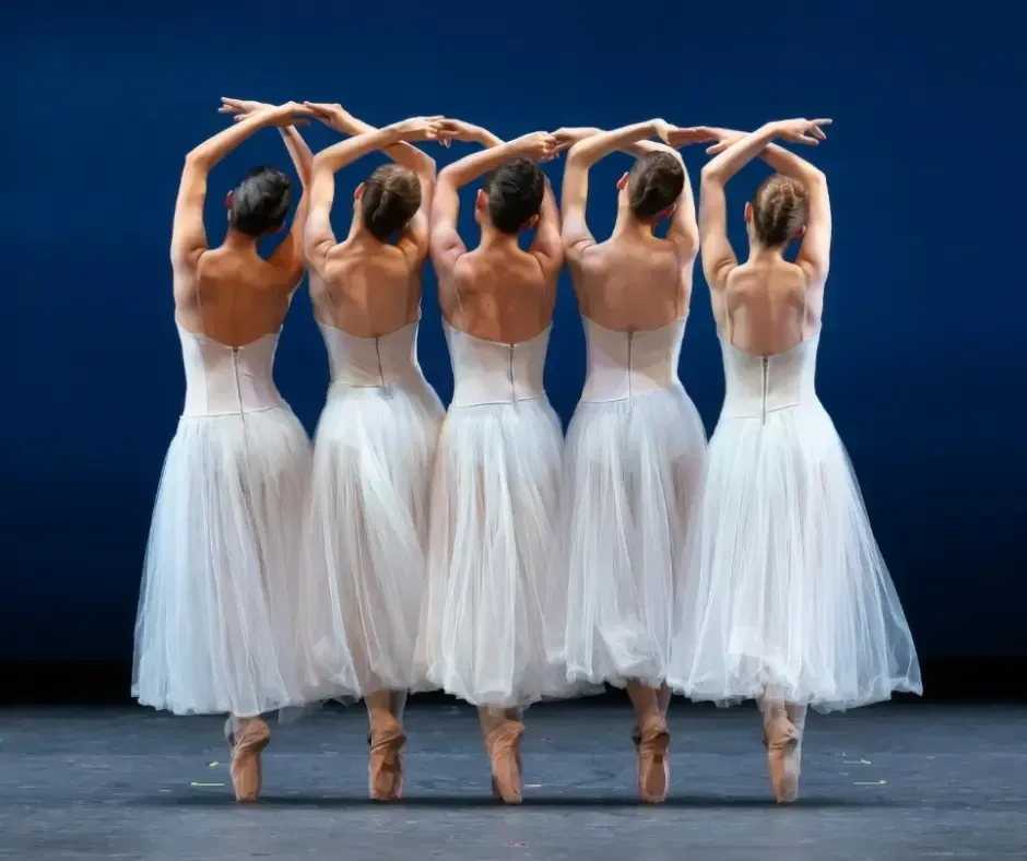 Five ballerinas in white tutus, arms arched overhead, on pointe, backs to viewer.