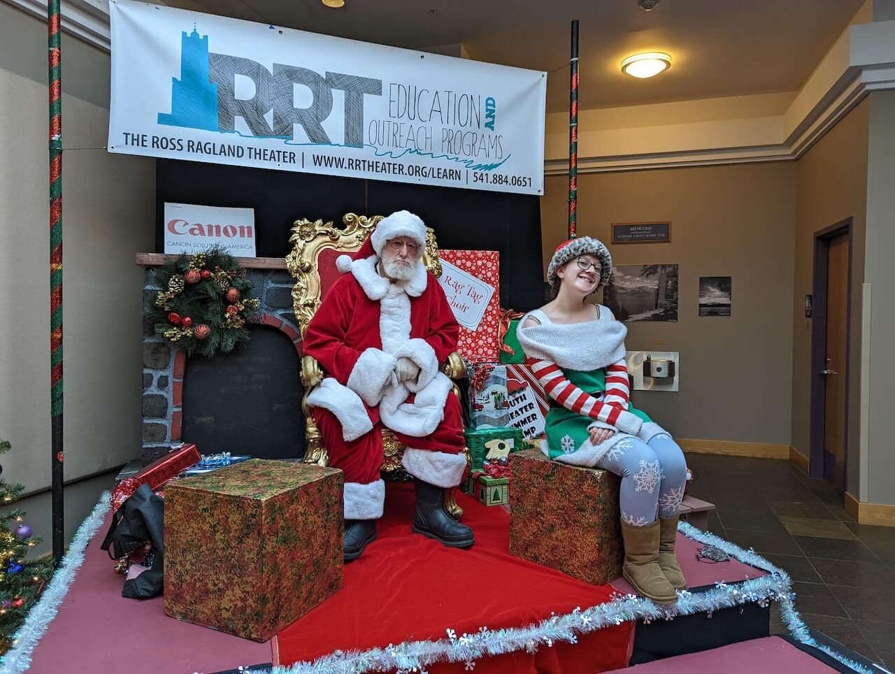 Santa Claus and an elf sit on a stage decorated with Christmas decor. A banner is above them.