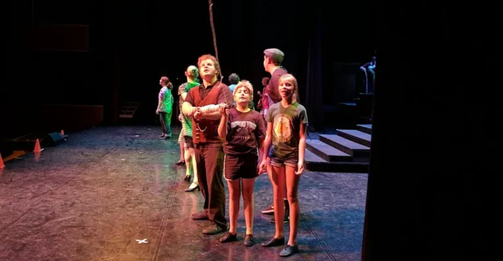 People on a stage looking up. Some wearing casual clothes, some in costumes. Dark stage setting.