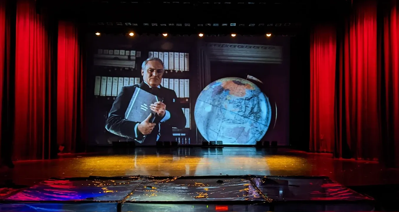 Stage with screen showing a man holding a book and a globe. Red curtains frame the scene.