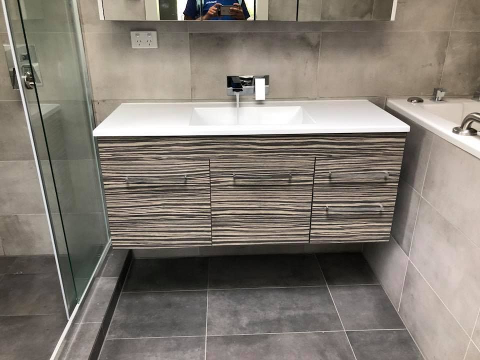 Floating bathroom vanity with white countertop, textured wood-grain cabinets, silver faucet, and grey tile. — Precise Plumbing & Drainage in Belconnen, ACT