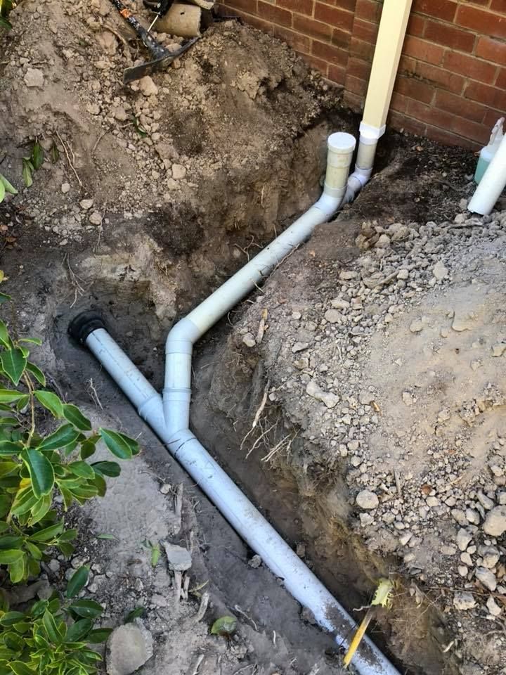 Excavated trench with PVC drain pipes extending from a downspout along a brick wall, in outdoor setting. — Precise Plumbing & Drainage in Belconnen, ACT