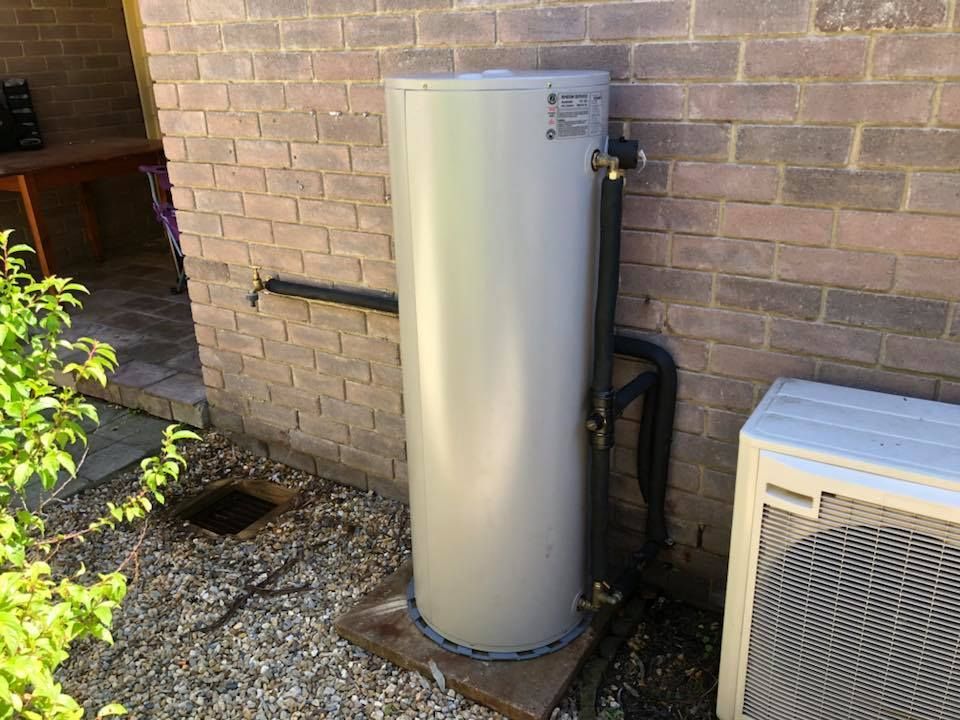 Water heater beside a brick wall; black pipes run along the side, next to an air conditioning unit. — Precise Plumbing & Drainage in Tuggeranong, ACT