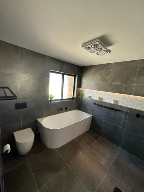 Modern bathroom with grey tiled walls and floor, white bathtub and toilet. — Precise Plumbing & Drainage In Conder, ACT