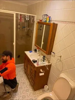 Man working in bathroom. Wooden vanity with mirror, toilet, shower, and tiled walls. — Precise Plumbing & Drainage in Tuggeranong, ACT