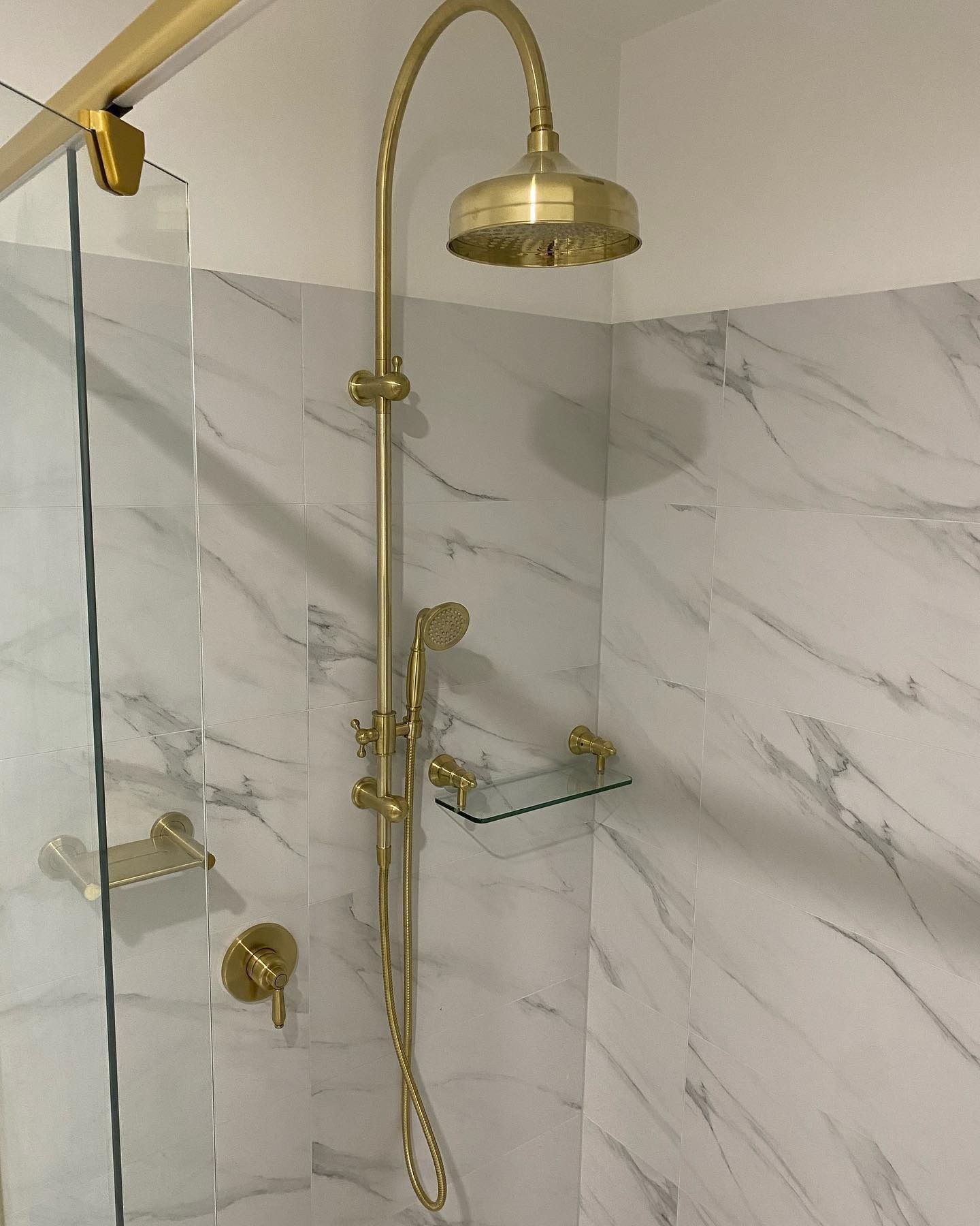 Gold shower fixture in a marble-tiled shower with a glass shelf and door. — Precise Plumbing & Drainage in Gungahlin, ACT